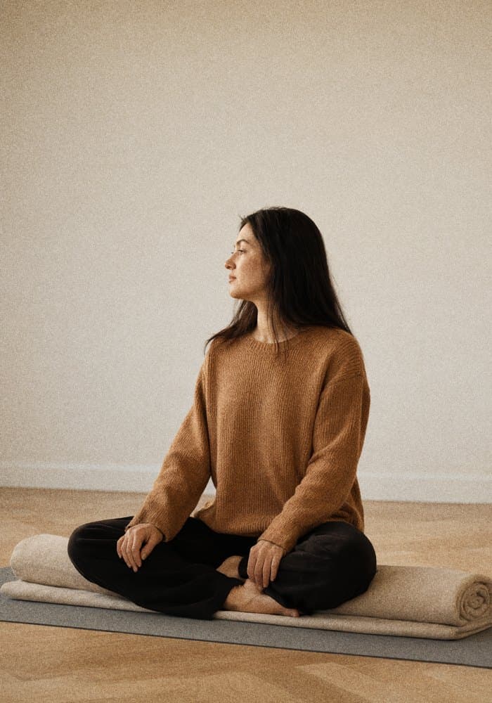 Porträt einer Yogalehrenden im Sitzen auf einer gefalteten Decke, Profil zum Fenster, gedämpftes Abendlicht.
